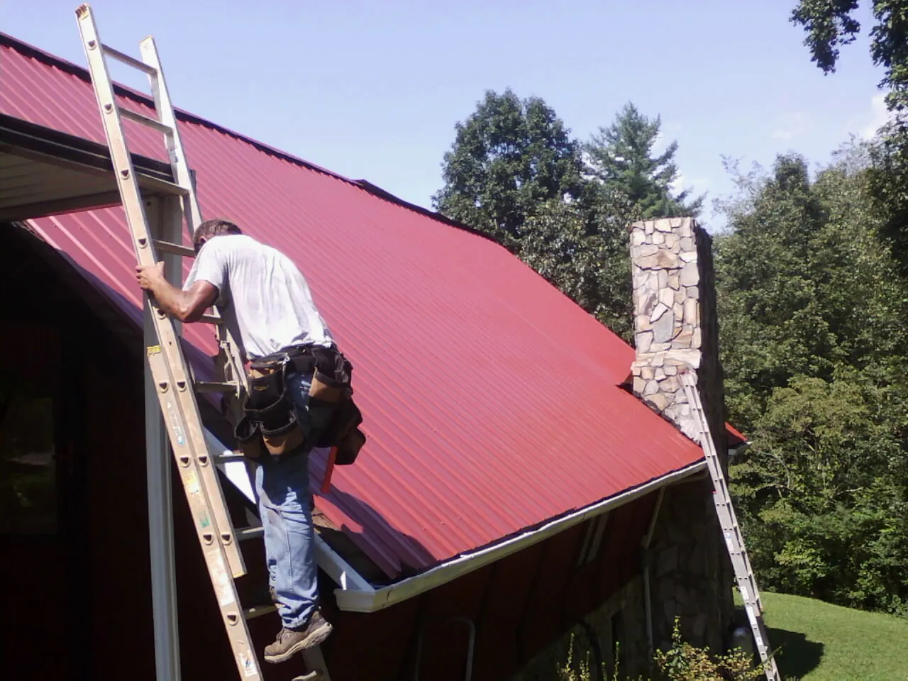 Roofer on a ladder beside a red metal roof and stone chimney — Soffit & Fascia Repair by Roofing Brothers in Evadale